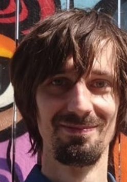 Fotografia młodego mężczyzny, Tomasza S. Markiewki, na tle kolorowego muralu w fantazyjne kształty. Markiewka ma dłuższe, brązowe włosy, zarost, uśmiecha się. Ubrany jest w niebieską, dżinsową koszulę, na plecach ma plecak.