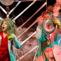 Na zdjęciu widać dwoje naukowców, kobietę i mężczyznę o siwych, bujnych włosach. Bohaterka Nielemka ubrana jest w czerwony kombinezon oraz biały fartuch opatrzony różnymi wzorami chemicznymi i matematycznymi. Bohater Lemtak ma widoczne na czole okulary i dmucha w rurę wentylacyjną, z której wylatuje dym.