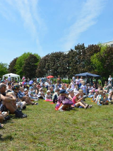 Grupa dzieci i dorosłych siedzących na trawie. W tle drzewa, niebo i blok.