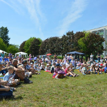 Grupa dzieci i dorosłych siedzących na trawie. W tle drzewa, niebo i blok.