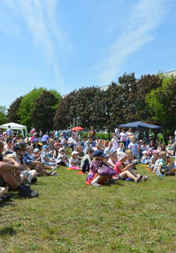 Grupa dzieci i dorosłych siedzących na trawie. W tle drzewa, niebo i blok.