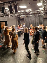 Grupa ludzi tańczy w kręgu na scenie. Prawa rękę mają wyciągnietą do środka.