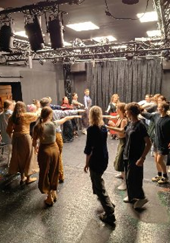 Grupa ludzi tańczy w kręgu na scenie. Prawa rękę mają wyciągnietą do środka.