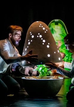 Dwoje ludzi otwiera świecącą kapsułę w której znajduje się roślinka. W tle można zaobserwować inne osoby które patrzą na całość z zaciekawieniem.