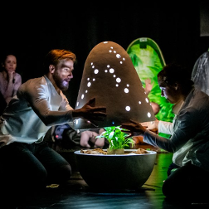 Dwoje ludzi otwiera świecącą kapsułę w której znajduje się roślinka. W tle inne osoby które patrzą z zaciekawieniem.