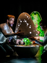 Dwoje ludzi otwiera świecącą kapsułę w której znajduje się roślinka. W tle można zaobserwować inne osoby które patrzą na całość z zaciekawieniem.