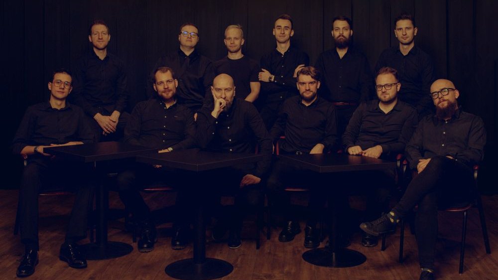 Mężczyżni w czarnych koszulach siedzą za stolikami barowymi, pozują do zdjęcia na ciemnym tle.