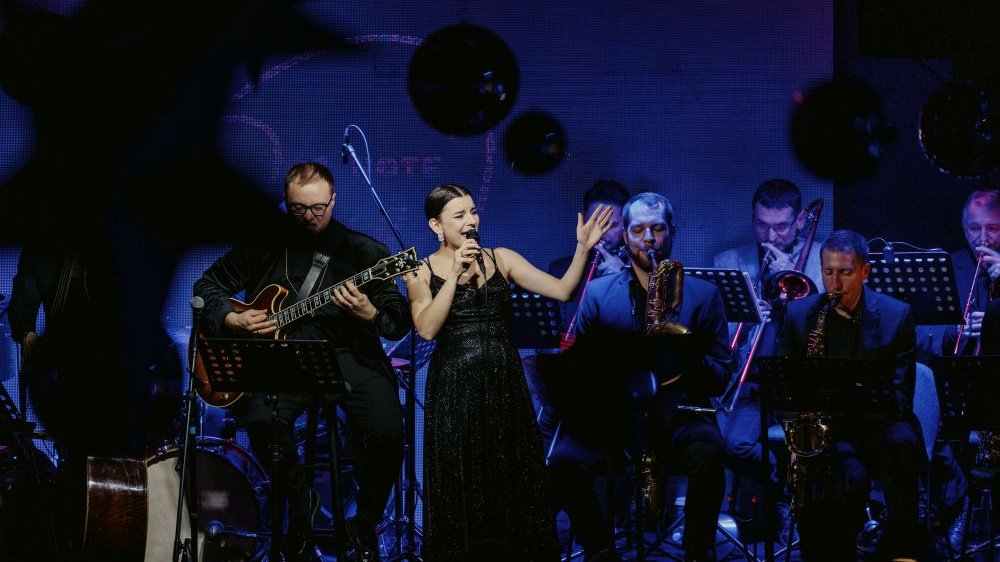 Oświetlona reflektorem wokalistka śpiewa na scenie. Za nią muzycy skąpani w niebieskim świetle.