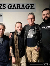 Kilku mężczyzn stoi w pomieszczeniu, w tle ściana, nad nimi napis informujący o nazwie zespołu i dacie koncertu.