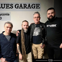 Kilku mężczyzn stoi w pomieszczeniu, w tle ściana, nad nimi napis informujący o nazwie zespołu i dacie koncertu.