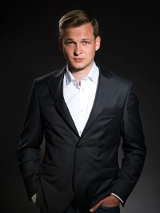 Na zdjęciu widoczny jest mężczyzna ubrany w elegancki czarny garnitur i białą koszulę. Stoi przed ciemnym tłem, które podkreśla jego sylwetkę. Mężczyzna ma krótkie jasne włosy i patrzy bezpośrednio w kamerę z lekkim uśmiechem. Ogólny ton fotografii tworzy wrażenie profesjonalizmu i pewności siebie.