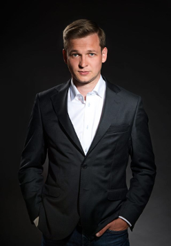 Na zdjęciu widoczny jest mężczyzna ubrany w elegancki czarny garnitur i białą koszulę. Stoi przed ciemnym tłem, które podkreśla jego sylwetkę. Mężczyzna ma krótkie jasne włosy i patrzy bezpośrednio w kamerę z lekkim uśmiechem. Ogólny ton fotografii tworzy wrażenie profesjonalizmu i pewności siebie.