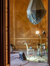 Przez otwarte drzwi widać fragment odnawianego pokoju. Na ścianach zabytkowa boazeria, żyrandol jest owinięty folią ochronną, a na podłodze stoją dwa krzesła, lampa i sporo kabli.