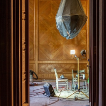 Przez otwarte drzwi widać fragment odnawianego pokoju. Na ścianach zabytkowa boazeria, żyrandol jest owinięty folią ochronną, a na podłodze stoją dwa krzesła, lampa i sporo kabli.