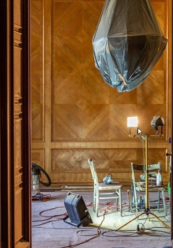 Przez otwarte drzwi widać fragment odnawianego pokoju. Na ścianach zabytkowa boazeria, żyrandol jest owinięty folią ochronną, a na podłodze stoją dwa krzesła, lampa i sporo kabli.