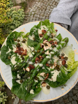 Zielone szparagi na liściu sałaty z roztopionym serem posypane nasionami i orzechami podane na białym talerzu.