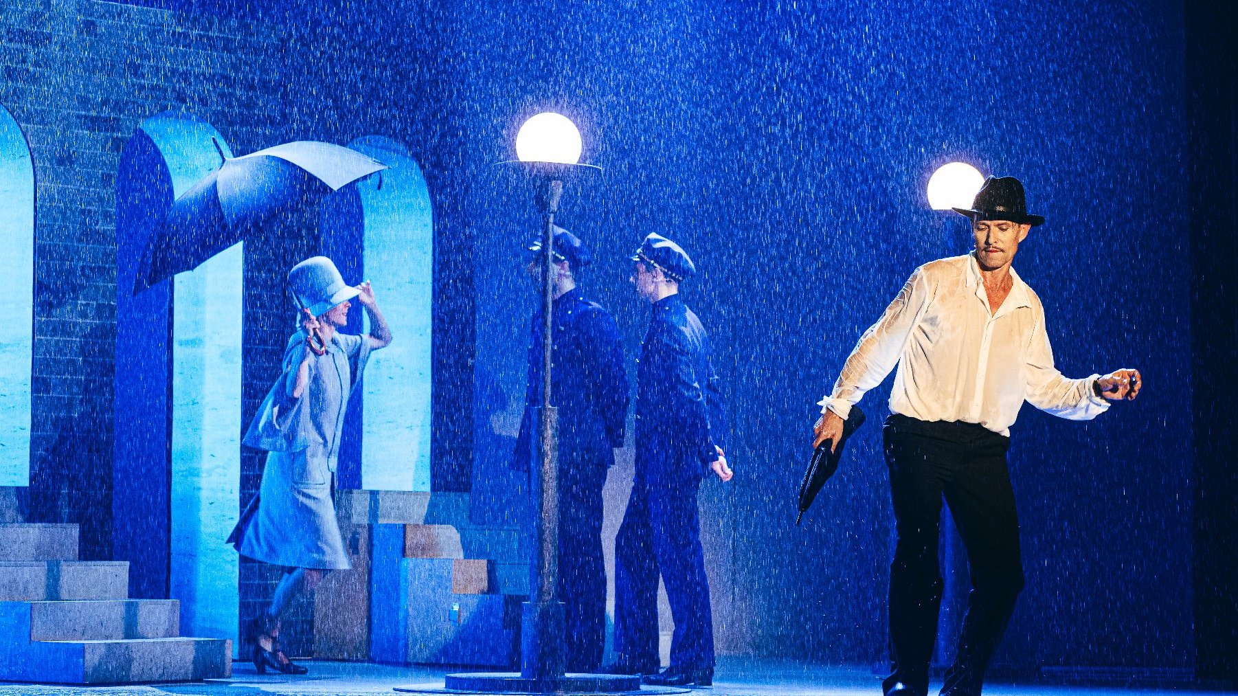 Scena z Deszczowej Piosenki w teatrze, aktor tańczy z parasolem, w tle policjanci, obok aktorka w kapeluszu z lat 20.