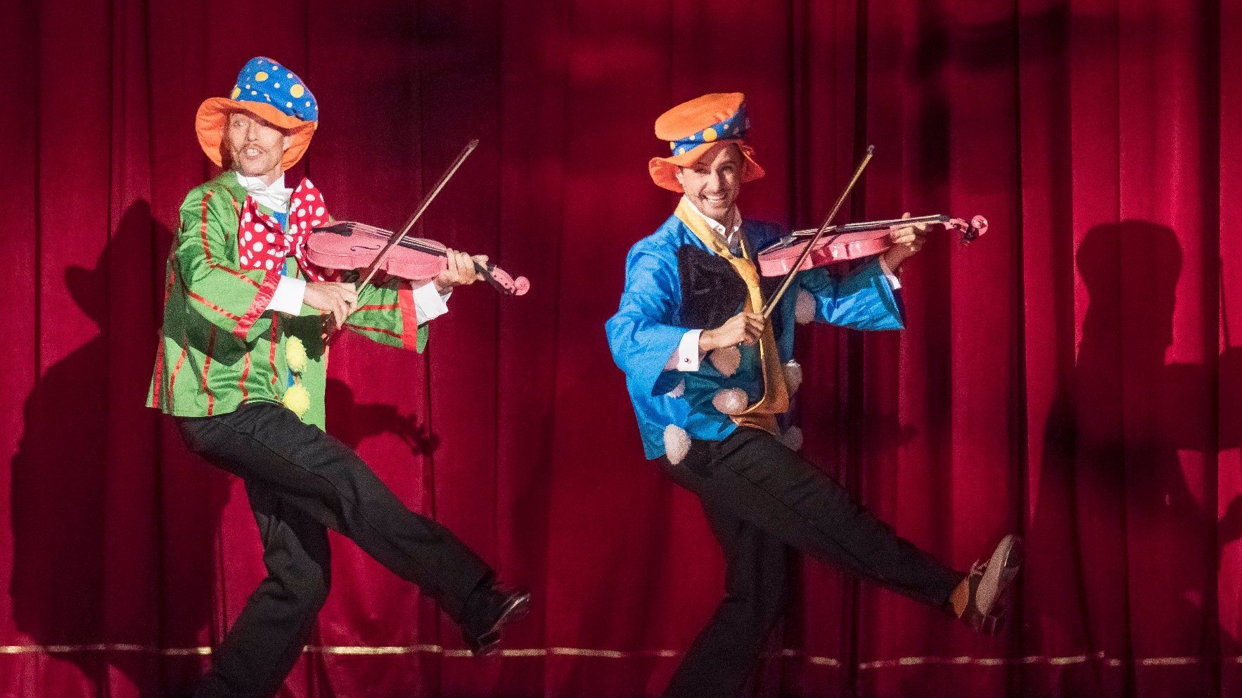 Dwóch kolorowo i fantazyjnie ubranych aktorów tańczy i śpiewa na scenie wspomagając się grą na różowych skrzypcach
