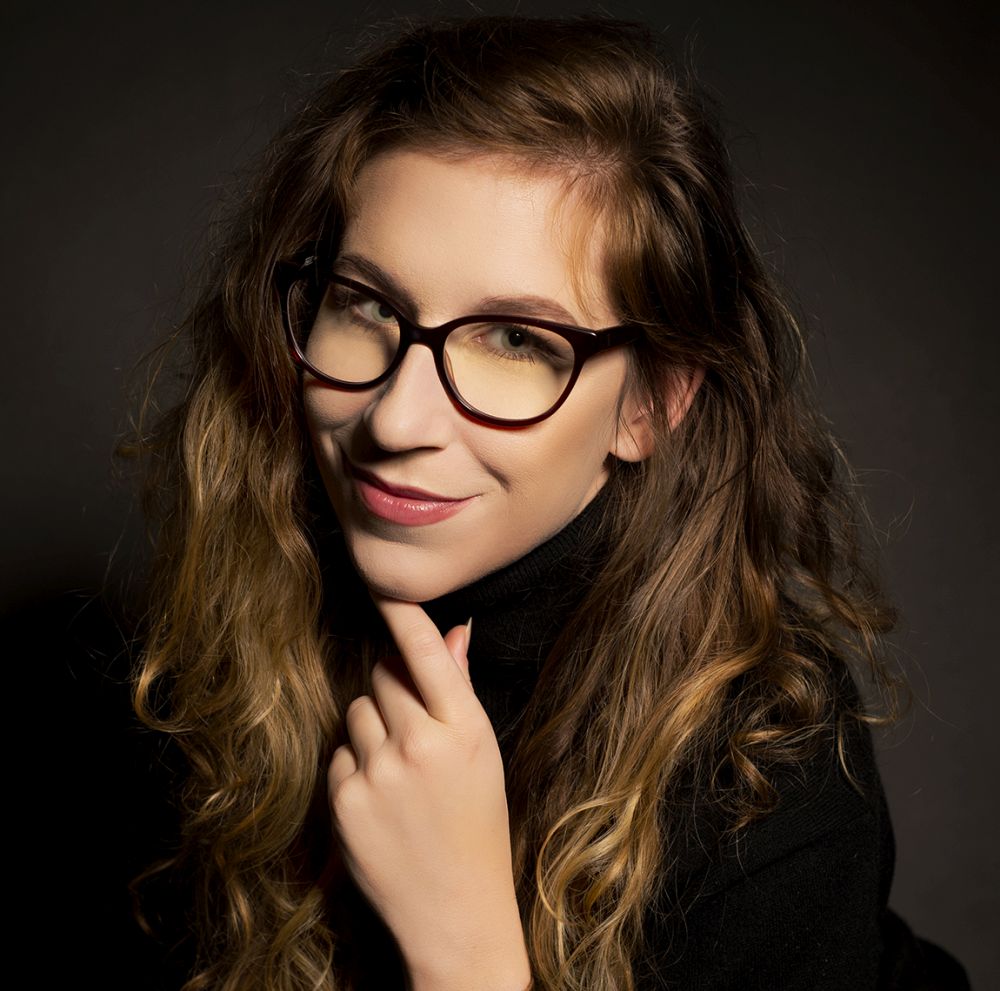 Magdalena Milert, fot. Adrianna Sołtys Długowłosa brunetka w okularach uśmiecha się podpierając brodę ręką.