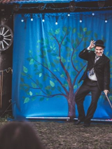 Mężczyzna z laską w dłoni i w meloniku stoi na scenie, za nim niebieska płachta z namalowanym drzewem.