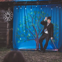 Mężczyzna z laską w dłoni i w meloniku stoi na scenie, za nim niebieska płachta z namalowanym drzewem.