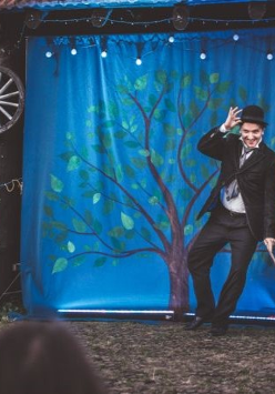 Mężczyzna z laską w dłoni i w meloniku stoi na scenie, za nim niebieska płachta z namalowanym drzewem.