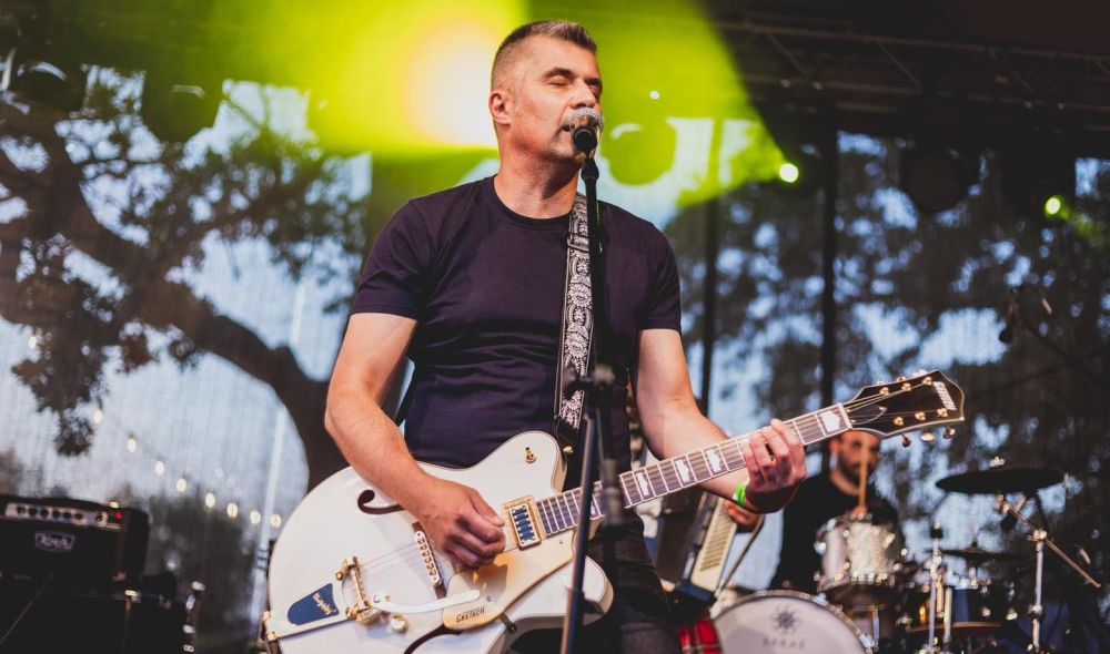 Dojrzały mężczyzna z gitarą elektryczną i przy mikrofonie. Stoi na scenie, ma zamknięte oczy.