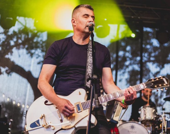 Dojrzały mężczyzna z gitarą elektryczną i przy mikrofonie. Stoi na scenie, ma zamknięte oczy.