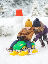 Dwójka dzieci bawiąca się w śniegu. Za nimi bałwanek i choinki pokryte śniegiem.