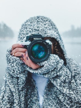 Osoba w czapce stoi z aparatem, w tle ośnieżone jezioro i las.
