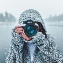 Osoba w czapce stoi z aparatem, w tle ośnieżone jezioro i las.