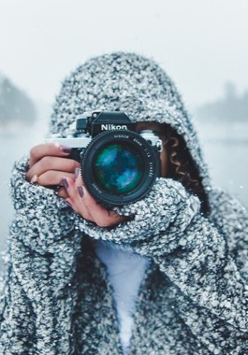 Osoba w czapce stoi z aparatem, w tle ośnieżone jezioro i las.