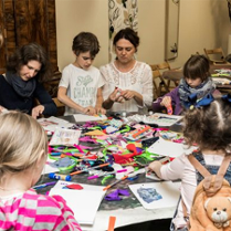 Opiekunowie z dziećmi na zajęciach przy stole pełnym kartek i kolorowych papierów.