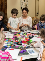 Dzieci z opiekunami na zajęciach przy stole pełnym kolorowych papierów.