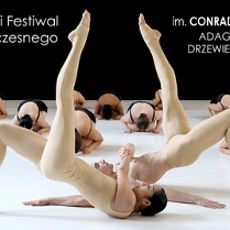 Obraz przedstawia grupę tancerzy występujących na scenie. Na pierwszym planie dwie tancerki znajdują się w efektownej pozie, jedna z nich balansuje na udach drugiej, z nogami wyciągniętymi w górę i splecionymi. Mają na sobie obcisłe, cieliste kostiumy, które zakrywają ich ciała od szyi w dół, sprawiając, że wydają się prawie nadzy i podkreślając linie ich mięśni oraz kształty tworzone przez ich pozy.