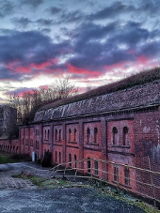 Budynki fortyfikacji z czerwonej cegły. Przy nich droga, a na niej samochód wojskowy.