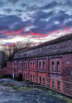 Budynki fortyfikacji z czerwonej cegły. Przy nich droga, a na niej samochód wojskowy.