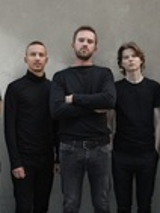 Fotografia przedstawia pięciu, ubranych na czarno mężczyzn, stojących na tle betonowej ściany. Mężczyzna znajdujący się w centrum i dwaj stojący na lewym i prawym skraju kadru mają założone ręce. Wszyscy patrzą prosto w obiektyw.