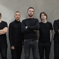 Fotografia przedstawia pięciu, ubranych na czarno mężczyzn, stojących na tle betonowej ściany. Mężczyzna znajdujący się w centrum i dwaj stojący na lewym i prawym skraju kadru mają założone ręce. Wszyscy patrzą prosto w obiektyw.