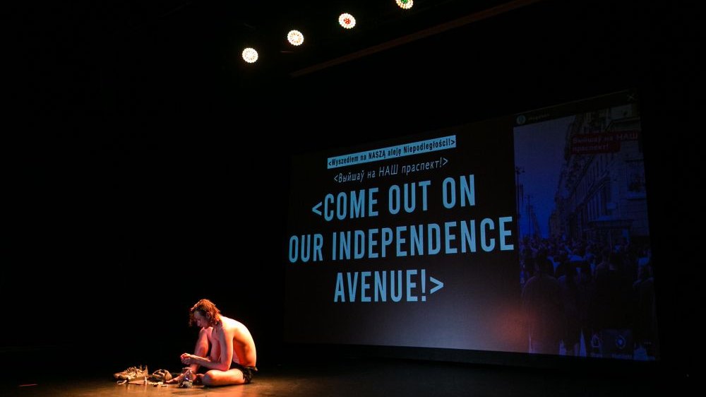 Ciemna sala. Performer siedzi na scenie otoczony przedmiotami. Za nim wyświetla się napis "Come out on our independence avenue!"