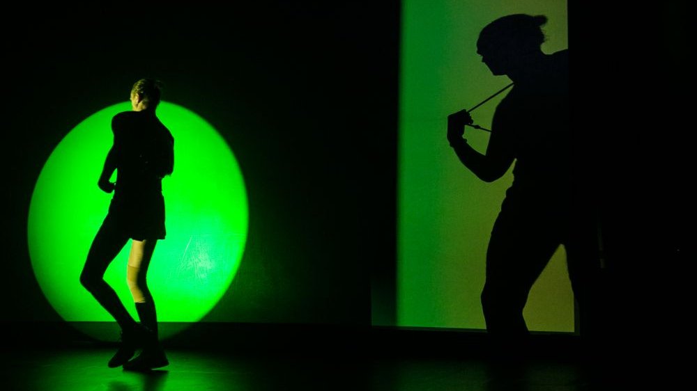 Perfomer idzie przez scenę, widać zielone światło, które odbija jego cień na ciemnej ścianie.