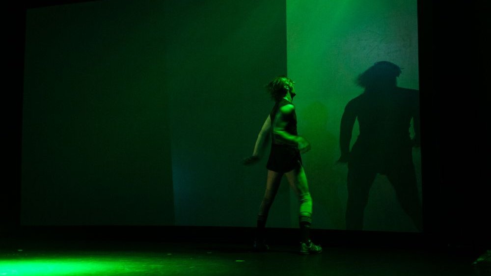 Podświetlony na zielono perfomer tańczy tyłem do publiczności, przodem do ściany, na której odbija się jego cień.