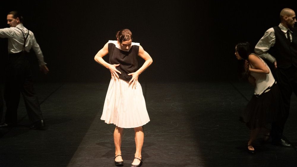 Kobieta stoi na środku sceny, trzyma się za brzuch. Wokół niej pozostali tancerze.