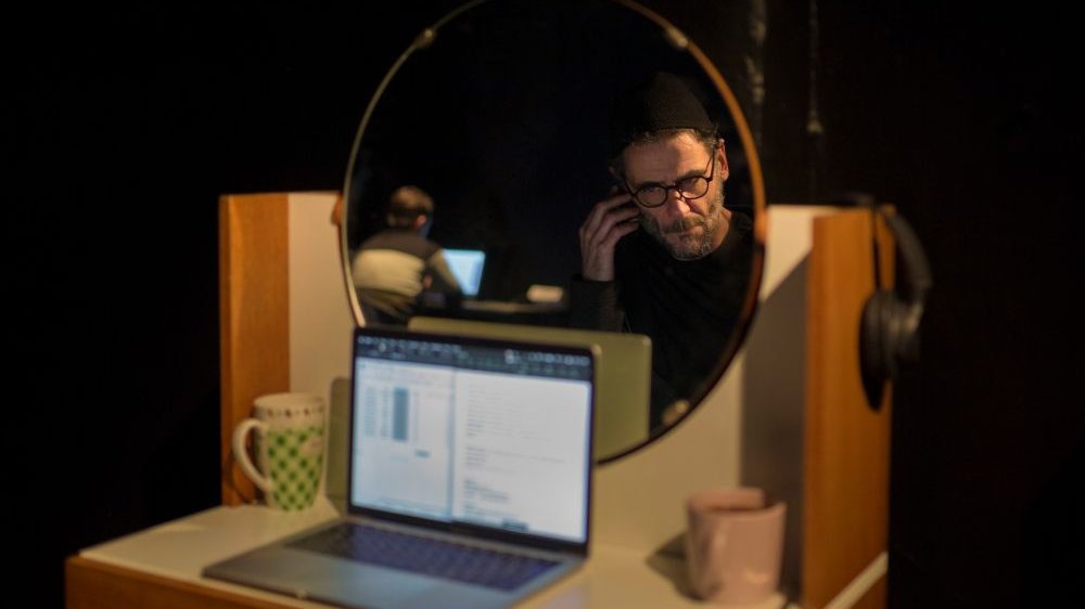 Mężczyzna w okularach oraz słuchawkach z mikrofonem odbity w lustrze. Na stoliku przed nim otwarty laptop i dwa kubki.