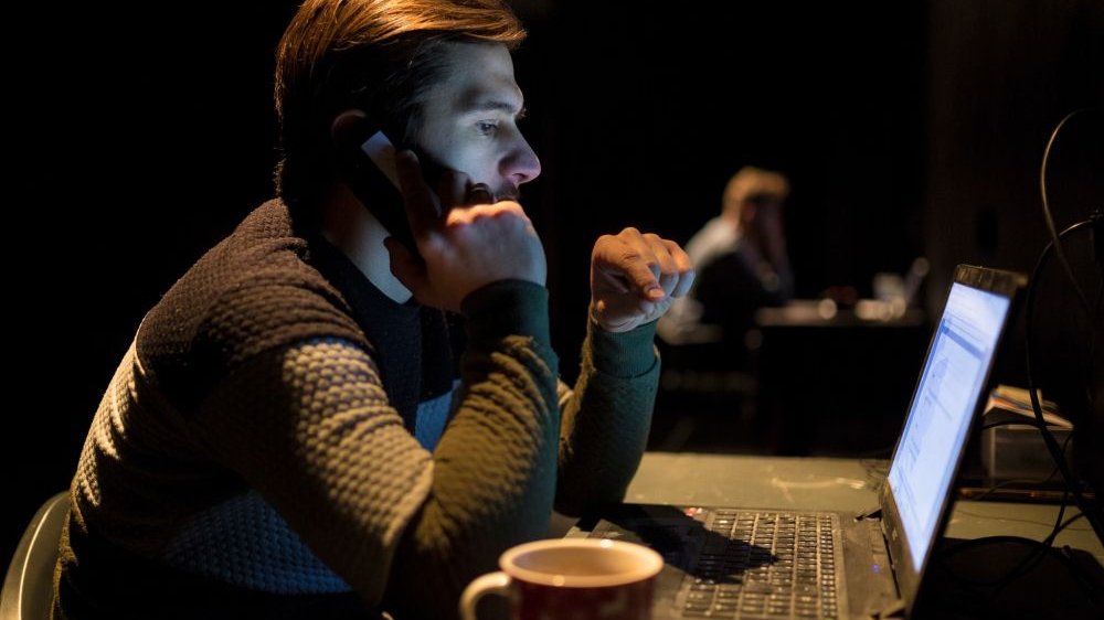 Mężczyzna siedzący przy biurku spogląda w ekran komputera, rozmawia przez telefon. Po jego prawej stronie stoi kubek.