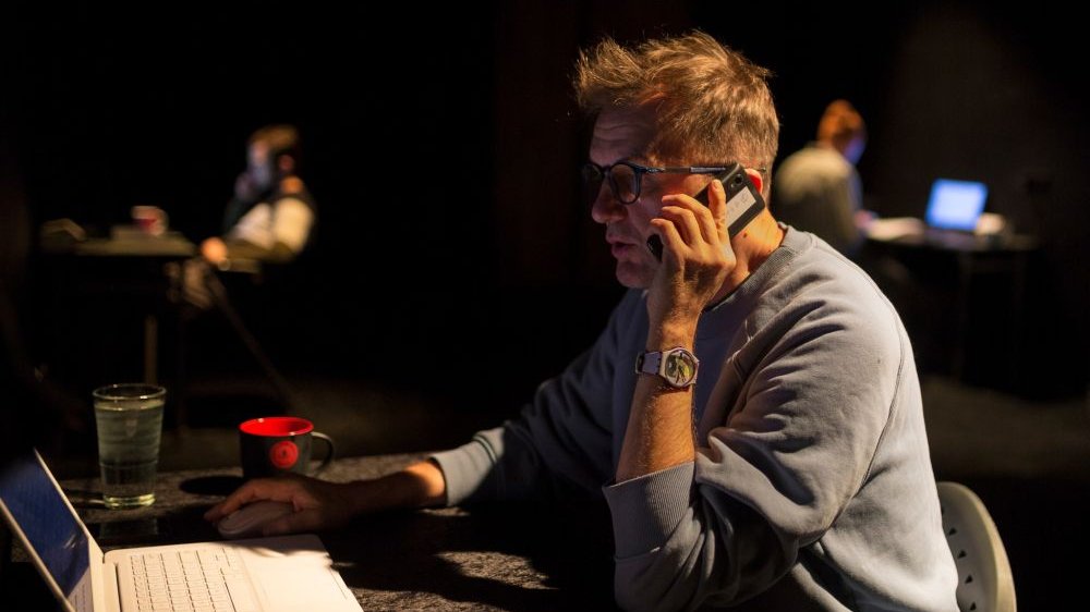 Mężczyzna rozmawia przez telefon, siedzi przy biurku. Na jego blacie biały laptop, szklanka i kubek.