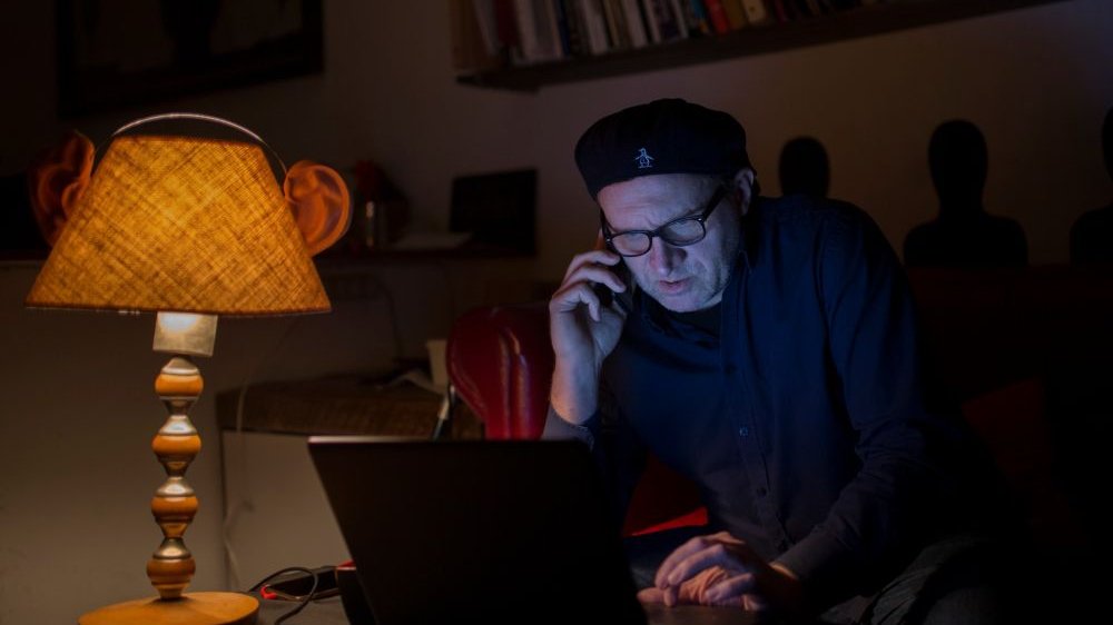Mężczyzna w kaszkiecie na głowie siedzi przed komputerem w półmroku, rozmawia przez telefon. Po jego prawej stronie nocna lampka, z tyłu półka z książkami.