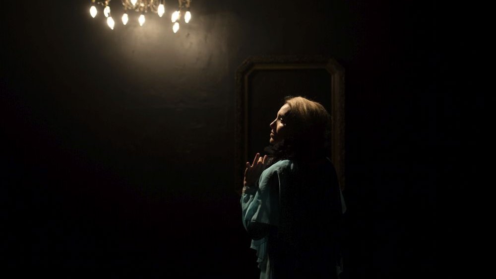 Kobieta stoi profilem do fotografa, unosi dłoń. Jest ciemno, pali się tylko jedna lampa górna z żyrandolem.