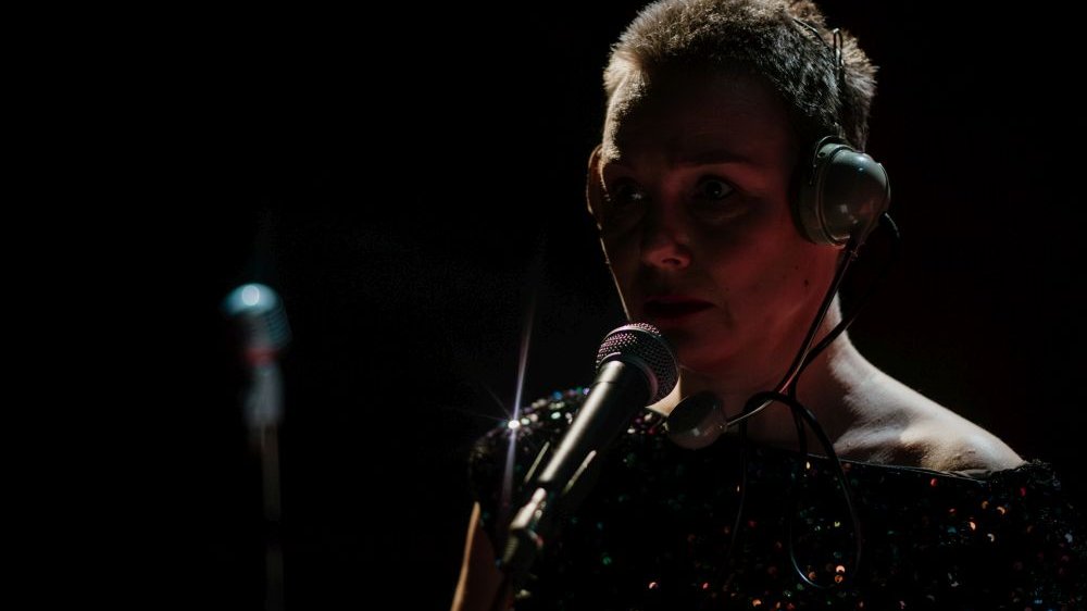 Aktorka w słuchawkach na głowie stoi przy mikrofonie w ciemności.