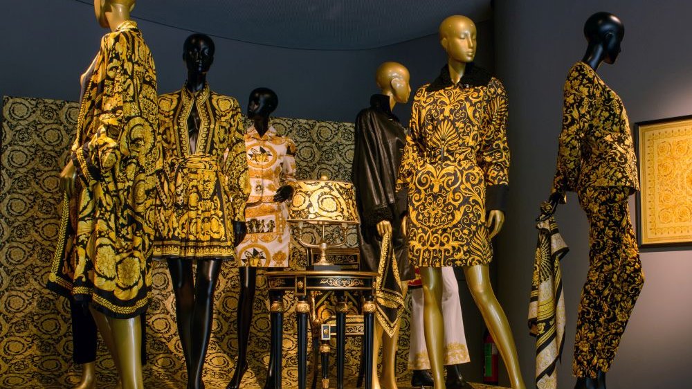 Złote i czarne kobiece manekiny prezentują ubrania - sukienki, garnitury i kimona w mocne, złote ornamenty. Za nimi ściana zdobiona w podobnych, złotych wzorach.